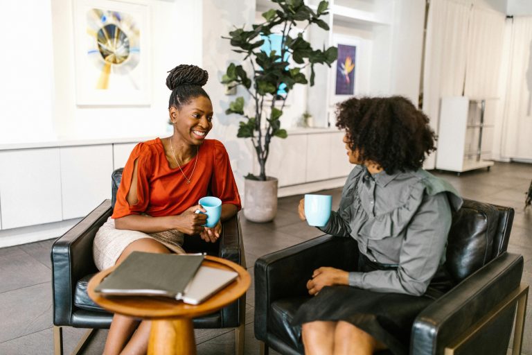 Two businesswomen enjoy a coffee break and conversation in a modern office setting.