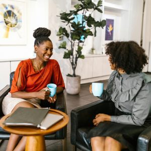 Two businesswomen enjoy a coffee break and conversation in a modern office setting.